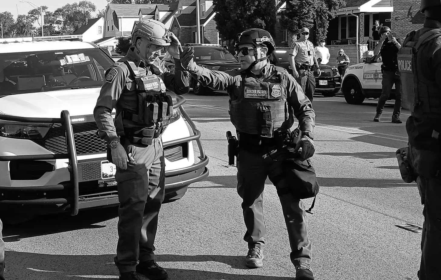 Black and white photo of two men in Bordal Patrol Uniforms standing on a road in a house-lined street. One is fixing the helmet of the other.