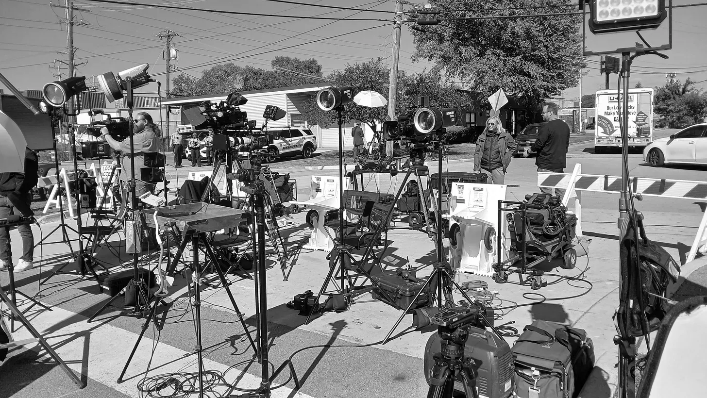 A black and white photo of lights and video cameras set up in the middle of a street next to barricades.